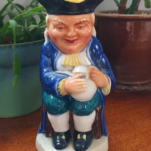Könnte beinhalten: Eine Keramikfigur eines Mannes, der einen blauen Mantel und einen Dreispitz trägt. Er sitzt auf einem Stuhl und hält eine Tasse. Die Figur ist in Blautönen, Weiß und Gelb bemalt.