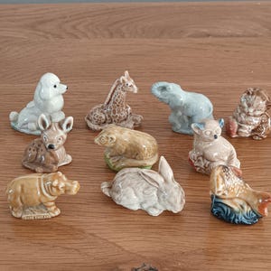 May include: A collection of small ceramic animal figurines, including a poodle, giraffe, elephant, cat, deer, bear, chihuahua, rabbit, hippo, and a bird. The figurines are in various colors and poses, set on a wooden surface.