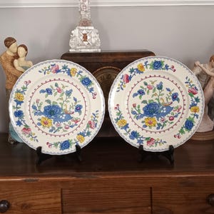 Puede incluir: Dos platos decorativos color crema con diseños florales. Cada plato presenta una flor azul central rodeada de acentos florales amarillos, rosas y azules. Los platos se exhiben sobre soportes negros, mostrando su intrincado diseño.