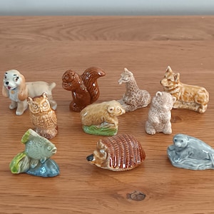 May include: A collection of ceramic animal figurines, including a dog, squirrel, owl, fish, bear, giraffe, hedgehog, corgi, and seal. The figurines are painted in various colors and sit on a wooden surface.