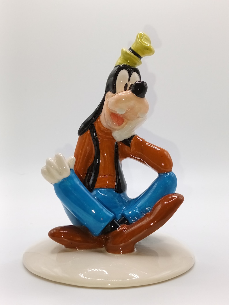 May include: A ceramic figurine of Goofy sitting with his legs crossed, wearing a blue shirt, brown trousers, and a yellow hat. He is smiling and has a white background.