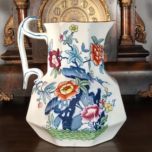 Puede incluir: Una jarra de cerámica blanca con un diseño floral con flores rojas, naranjas, azules y rosas. La jarra tiene un asa única y un borde decorativo. Es una pieza vintage.