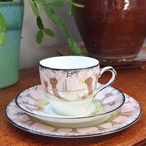 Op de afbeelding: Een delicate theekop en schotelset met een wilgenboomontwerp. De kop en schotel zijn wit met een zwarte rand en een roze achtergrond. De wilgenbomen zijn geschilderd in bruin en groen.