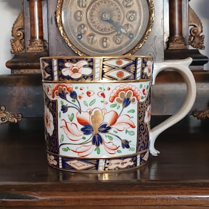May include: A ceramic mug featuring a floral pattern in blue, orange, and white, with gold detailing. The mug has a curved handle and is displayed on a dark wooden surface. The mug's design is reminiscent of antique china.