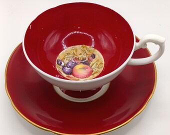 May include: A red teacup and saucer set with a white floral design featuring a basket of fruit. The cup has a white interior and a gold rim. The saucer has a scalloped edge and a gold rim.