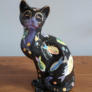 May include: Black ceramic cat figurine with gold accents and colorful fish designs. The cat is sitting upright with its tail curled. The fish are in shades of blue, yellow, and green. The cat's eyes and ears are outlined in gold.