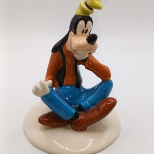 May include: A ceramic figurine of Goofy sitting with his legs crossed, wearing a brown shirt, blue jeans, and a yellow hat. He is smiling and has a white glove on his left hand.