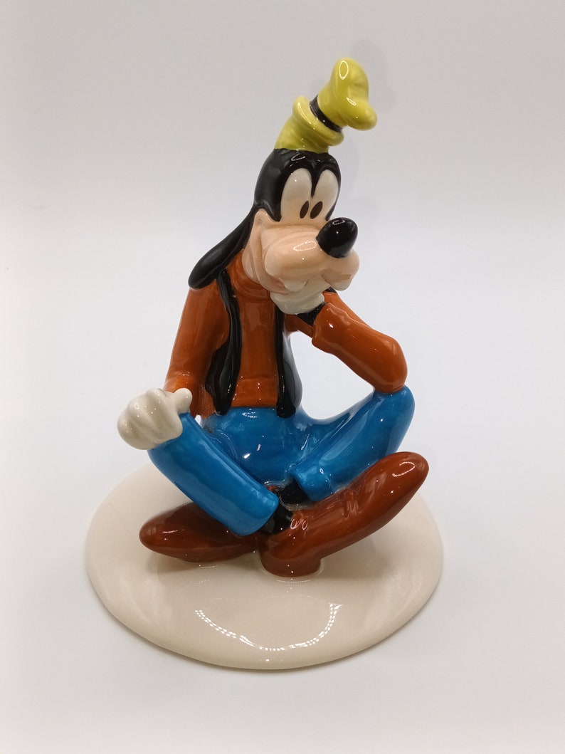 May include: A ceramic figurine of Goofy sitting with his legs crossed, wearing a brown shirt, blue jeans, and a yellow hat. He is smiling and has a white glove on his left hand.
