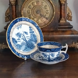 Op de afbeelding: Een blauw-wit porseleinen theeservies met een Willow-patroon. De set bevat een theekopje, een schotel en een klein bord, allemaal met gouden randen. De items worden gepresenteerd op een donker houten oppervlak, met een antieke klok op de achtergrond.