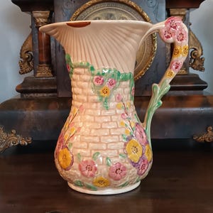 Peut inclure: Pichet décoratif en céramique avec un dessus crème en forme d'éventail et un corps en forme de briques. Le pichet est orné de motifs floraux colorés roses, jaunes et violets. La poignée est décorée de fleurs roses et de feuilles vertes.