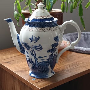 Puede incluir: Una tetera de porcelana blanca con un diseño de paisaje azul y blanco. La tetera tiene un pico curvo, un asa de bucle y una tapa con un adorno. El diseño presenta pájaros, árboles y edificios.