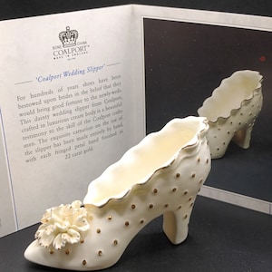 Op de afbeelding: Een witte porseleinen bruidsschoen met gouden accenten en een witte bloem op de teen. De schoen is versierd met gouden stippen en heeft een gouden rand rond de rand. 'Coalport Wedding Slipper' staat op de kaart.