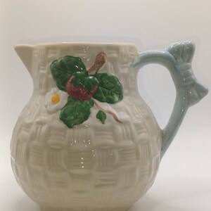 Peut inclure: Un pichet en céramique blanche avec un motif tissé et une poignée bleue. Le pichet présente un motif peint d'une fraise et de feuilles.