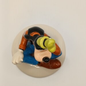 May include: A ceramic figurine of Goofy sitting down, wearing his trademark yellow hat, blue trousers, and brown shoes. The figurine is painted in bright colours and has a detailed design.