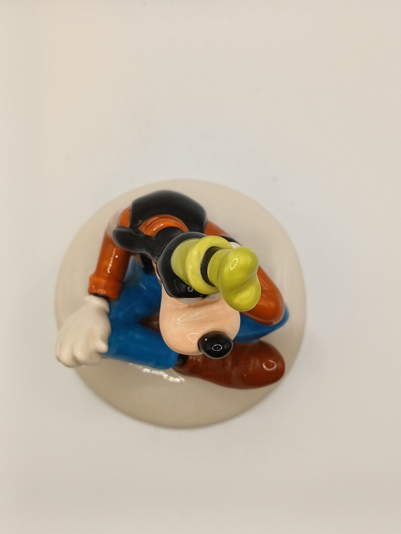 May include: A ceramic figurine of Goofy sitting down, wearing his trademark yellow hat, blue trousers, and brown shoes. The figurine is painted in bright colours and has a detailed design.