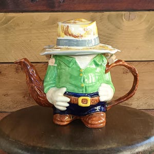 May include: A whimsical ceramic teapot shaped like a farmer. The teapot features a green shirt, blue pants, brown boots, and a straw hat. The handle and spout are brown, and the teapot is set on a wooden surface.