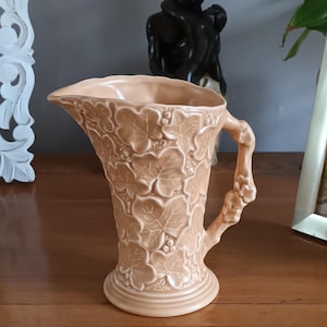 Może przedstawiać: Brzoskwiniowy ceramiczny dzban z wzorem liści i jagód. Dzban ma rozkloszowaną górę, zakrzywiony uchwyt i okrągłą podstawę. Wzór jest wytłoczony na powierzchni, dodając tekstury. Dzban prawdopodobnie do użytku dekoracyjnego.