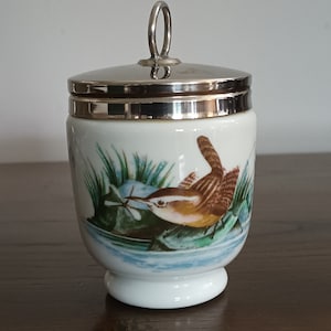 May include: White ceramic egg cup with a silver-colored lid and handle. The cup features a detailed illustration of a brown bird with a long beak, surrounded by green and blue foliage. A classic kitchenware item.