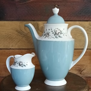 May include: A vintage tea set featuring a light blue and white color scheme. The set includes a tall teapot with a lid and a small creamer. Both pieces have a floral design around the top and are set on a wooden surface.