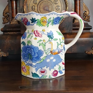 Könnte beinhalten: Ein weißer Keramikkrug mit gewelltem Rand und Henkel, verziert mit einem bunten Blumenmuster in Blau, Gelb, Rosa und Grün sowie einer stilisierten Vogelabbildung. Der Krug hat eine sechseckige Basis und eine sechseckige Körperform.
