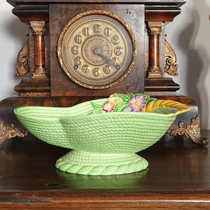 Puede incluir: Un cuenco de cerámica verde claro en forma de cornucopia con una superficie texturizada, apoyado sobre una base de pedestal. El cuenco está decorado con flores y hojas de colores. Un reloj antiguo está en el fondo.