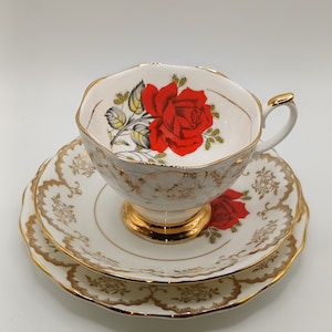 Pode incluir: Um conjunto de chávena e pires de chá branco com guarnição dourada e um desenho de rosa vermelha. A chávena de chá tem uma borda ondulada e o pires tem um padrão floral.