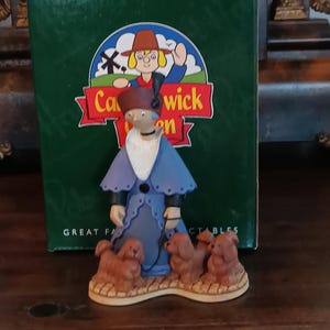 Könnte beinhalten: Eine Keramikfigur einer Frau in blauem Mantel und Hut, die eine Leine mit drei braunen Hunden hält. Die Figur steht vor einer grünen Schachtel mit der Aufschrift "Catwick Garden" und einer Cartoon-Illustration.