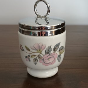 May include: A white ceramic egg cup with a silver lid and a floral design featuring pink roses and green leaves. The egg cup has a small handle on the lid.