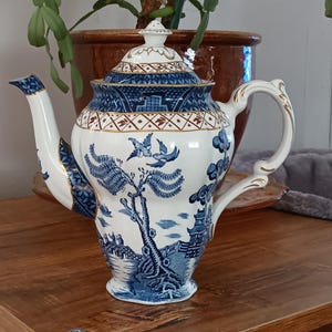 Könnte beinhalten: Eine weiße und blaue Keramik-Teekanne mit Deckel und detailliertem Landschaftsdesign. Die Teekanne hat einen gebogenen Ausguss und Griff mit goldenen Akzenten. Das Design umfasst Vögel, Bäume und Gebäude. Die Teekanne steht auf einer Holzoberfläche.