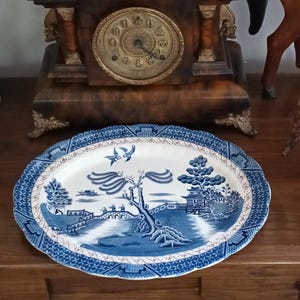 May include: Oval blue and white ceramic serving platter with a detailed Willow pattern design. The platter has a scalloped edge and a classic landscape scene with a bridge, trees, and birds. A decorative piece for serving.