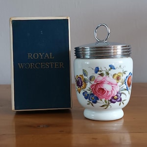 Op de afbeelding: Een witte Royal Worcester eierdop met een bloemmotief met roze rozen, blauwe en gele bloemen en paarse accenten. De eierdop heeft een zilverkleurig deksel en handvat. Een marineblauwe doos met "ROYAL WORCESTER" staat ernaast.