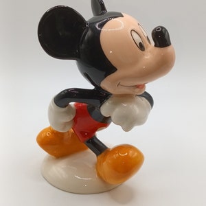 May include: A ceramic figurine of Mickey Mouse in a walking pose. He is wearing his signature red shorts, yellow shoes, and white gloves. His ears are black and his face is a light peach colour.