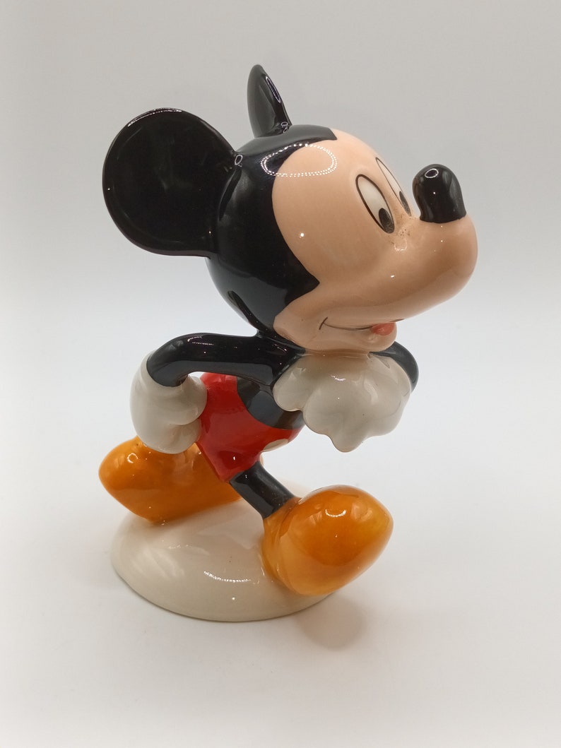 May include: A ceramic figurine of Mickey Mouse in a walking pose. He is wearing his signature red shorts, yellow shoes, and white gloves. His ears are black and his face is a light peach colour.