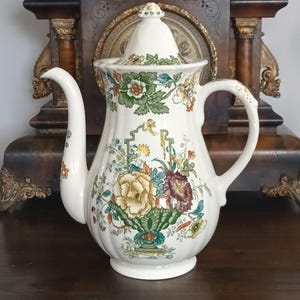 Puede incluir: Cafetera de cerámica blanca con un diseño floral en amarillo, verde y burdeos. La cafetera tiene un pico curvo, un asa y una tapa con un pequeño pomo. Un objeto de decoración para la mesa.