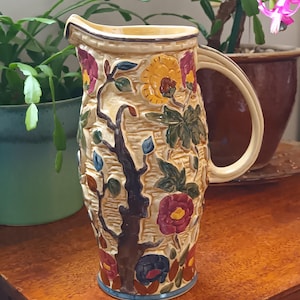 Puede incluir: Una jarra de cerámica con un diseño floral. La jarra es de color crema con flores y hojas marrones, verdes, rojas, amarillas y azules. La jarra tiene un asa y una boca ancha.