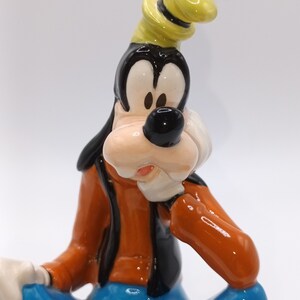 May include: A ceramic figurine of Goofy, a cartoon dog character from Disney, wearing a yellow hat and blue jeans. He is sitting with his right hand on his chin, looking thoughtful.