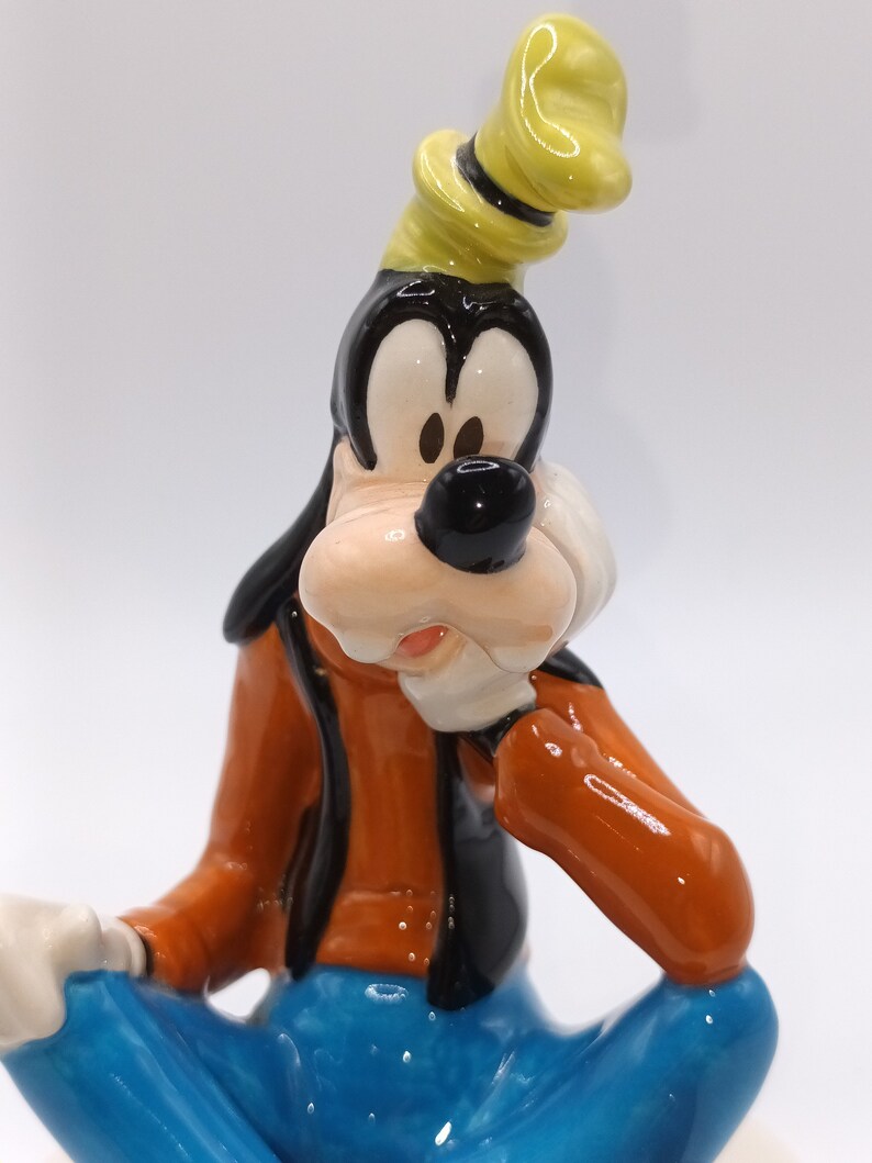May include: A ceramic figurine of Goofy, a cartoon dog character from Disney, wearing a yellow hat and blue jeans. He is sitting with his right hand on his chin, looking thoughtful.
