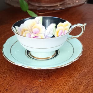 May include: A vintage teacup and saucer set featuring a floral design. The teacup has a black rim and a white interior with a floral pattern in shades of yellow, pink, and green. The saucer is a pale green colour with a gold rim.