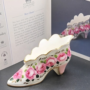 Op de afbeelding: Coalport porseleinen schoenfiguur, met het "Rose Cluster" patroon. De schoen is wit met roze rozen, groene bladeren en gouden randen. De hak is zachtroze. Het beeldje wordt gepresenteerd met een kaart met tekst.