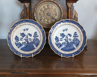May include: Two matching blue and white decorative plates with a gold rim and scalloped edges. The plates feature a classic Willow pattern design with a landscape scene. The plates are displayed on a wooden surface with an antique clock in the background.
