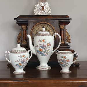 May include: A white porcelain tea set with a floral and bird design. The set includes a teapot, a lidded sugar bowl, and a creamer. The design features colourful birds and flowers. The set is displayed on a dark wooden surface.