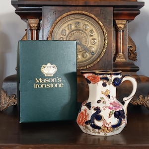 May include: A vintage white floral pitcher with a gold rim, featuring orange, pink, and blue floral designs. Beside it is a green box with the text "Mason's Ironstone" in gold lettering. The background includes a dark wooden clock.