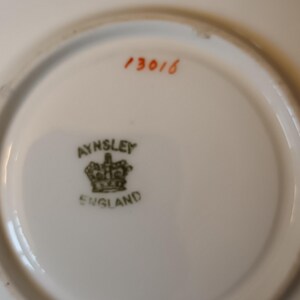 May include: The bottom of a white saucer with a green and gold Aynsley England logo and the number 13016 in orange.