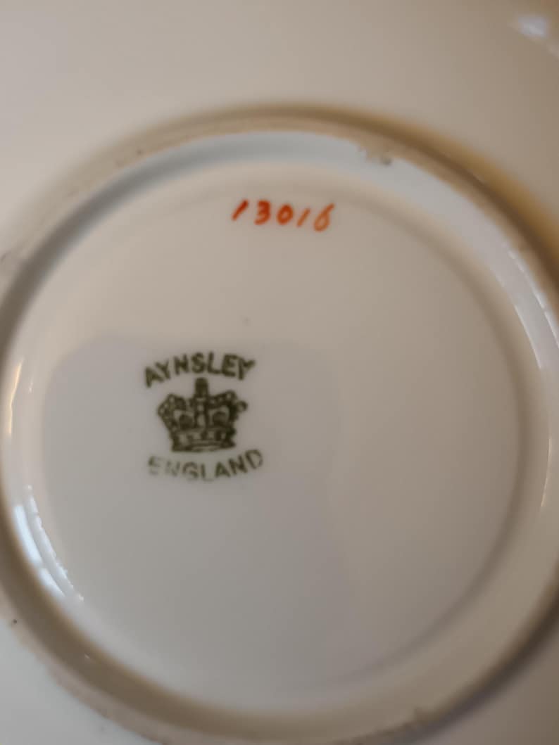 May include: The bottom of a white saucer with a green and gold Aynsley England logo and the number 13016 in orange.
