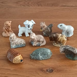 May include: A collection of small ceramic animal figurines, including a dog, squirrel, elephant, bear, and hedgehog. The figurines are in various colors, such as brown, white, and gray, and are arranged on a wooden surface.