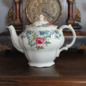 以下が含まれることがあります： 赤、青、黄色の花柄が施された白い磁器のティーポット。 ティーポットは、曲がった注ぎ口、金色のアクセントが付いたハンドル、お揃いの蓋が付いています。 背景にはヴィンテージの時計があります。
