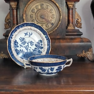 May include: A blue and white porcelain teacup and saucer set with a gold trim. The set features a detailed landscape design. A vintage clock is in the background.