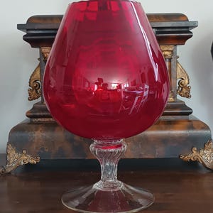 May include: A large, deep red glass vessel shaped like a brandy snifter, featuring a wide, rounded bowl with a scalloped rim. The bowl sits atop a clear, twisted glass stem that connects to a wide, circular, transparent glass base.