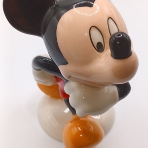 May include: A ceramic figurine of Mickey Mouse in a standing pose. Mickey is wearing his signature red shorts and yellow shoes. He has a black and white face with large, round eyes and a wide smile.