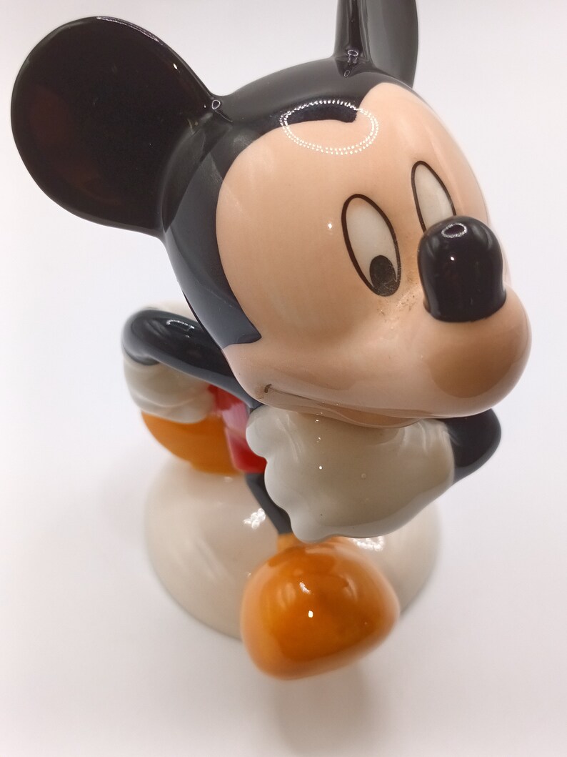 May include: A ceramic figurine of Mickey Mouse in a standing pose. Mickey is wearing his signature red shorts and yellow shoes. He has a black and white face with large, round eyes and a wide smile.